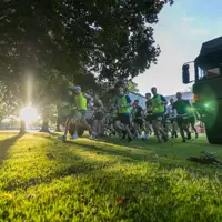 Runners in bright gear sprint across a sunlit park under large trees. The setting sun creates dramatic shadows; a truck is parked nearby. Energetic atmosphere.