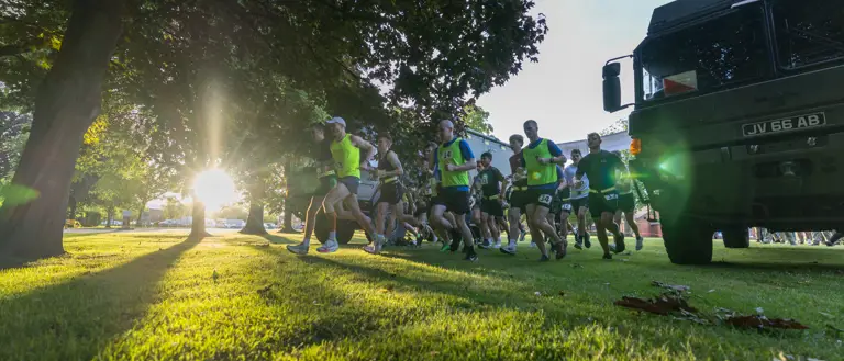 Runners in bright gear sprint across a sunlit park under large trees. The setting sun creates dramatic shadows; a truck is parked nearby. Energetic atmosphere.