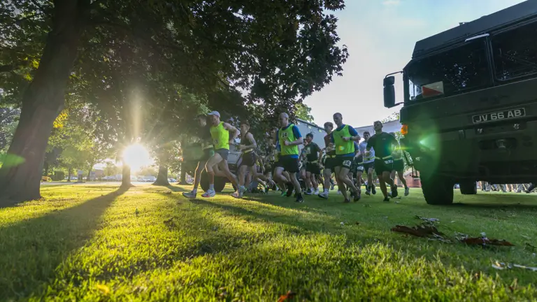 Runners in bright gear sprint across a sunlit park under large trees. The setting sun creates dramatic shadows; a truck is parked nearby. Energetic atmosphere.