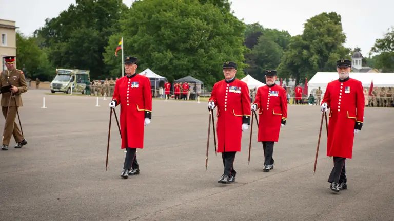 Four Royal Hospital Chelsea pensioners wearing long red dress tunics are marching across a parade square using pace sticks. Alongside them is an Army soldiers watching the team of four.