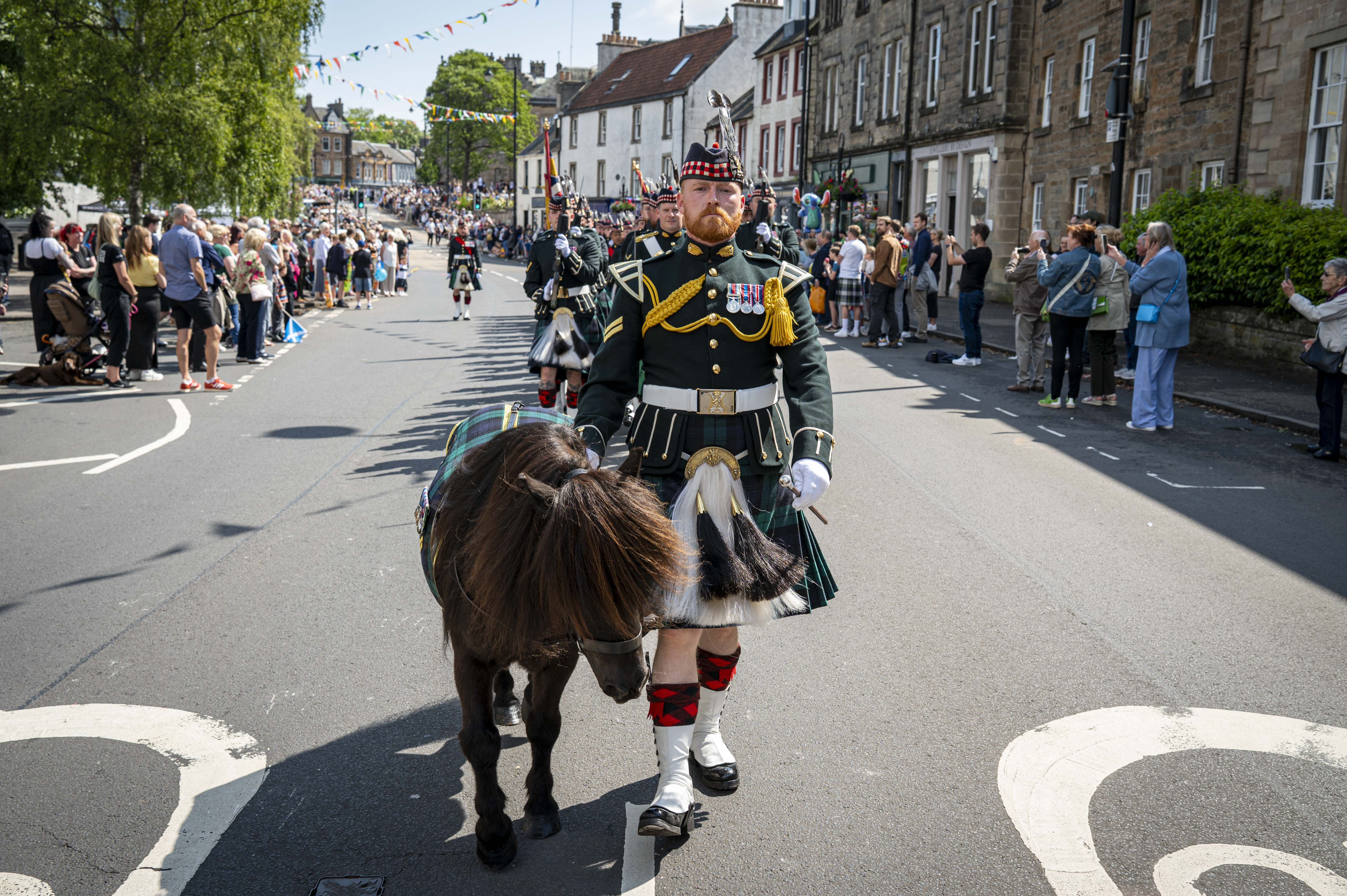 A man wearing a black tunic and a green and blue kilt marches in front of other soldiers. He is holding the reins of a Shetland Pony.