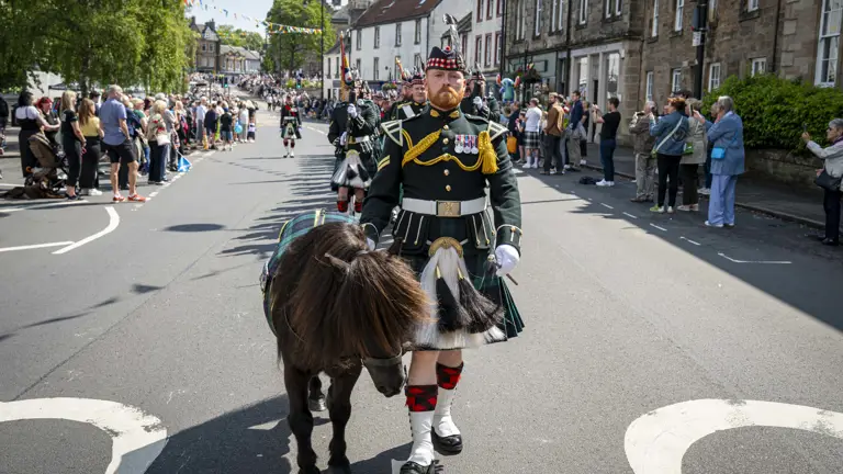 A man wearing a black tunic and a green and blue kilt marches in front of other soldiers. He is holding the reins of a Shetland Pony.