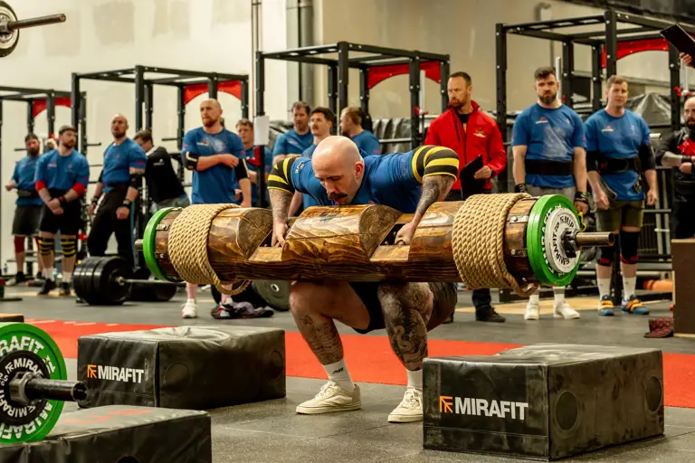 A bald man in a blue shirt and lifting straps squats with a large log on his shoulders, surrounded by onlookers in a gym setting.