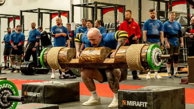 A bald man in a blue shirt and lifting straps squats with a large log on his shoulders, surrounded by onlookers in a gym setting.