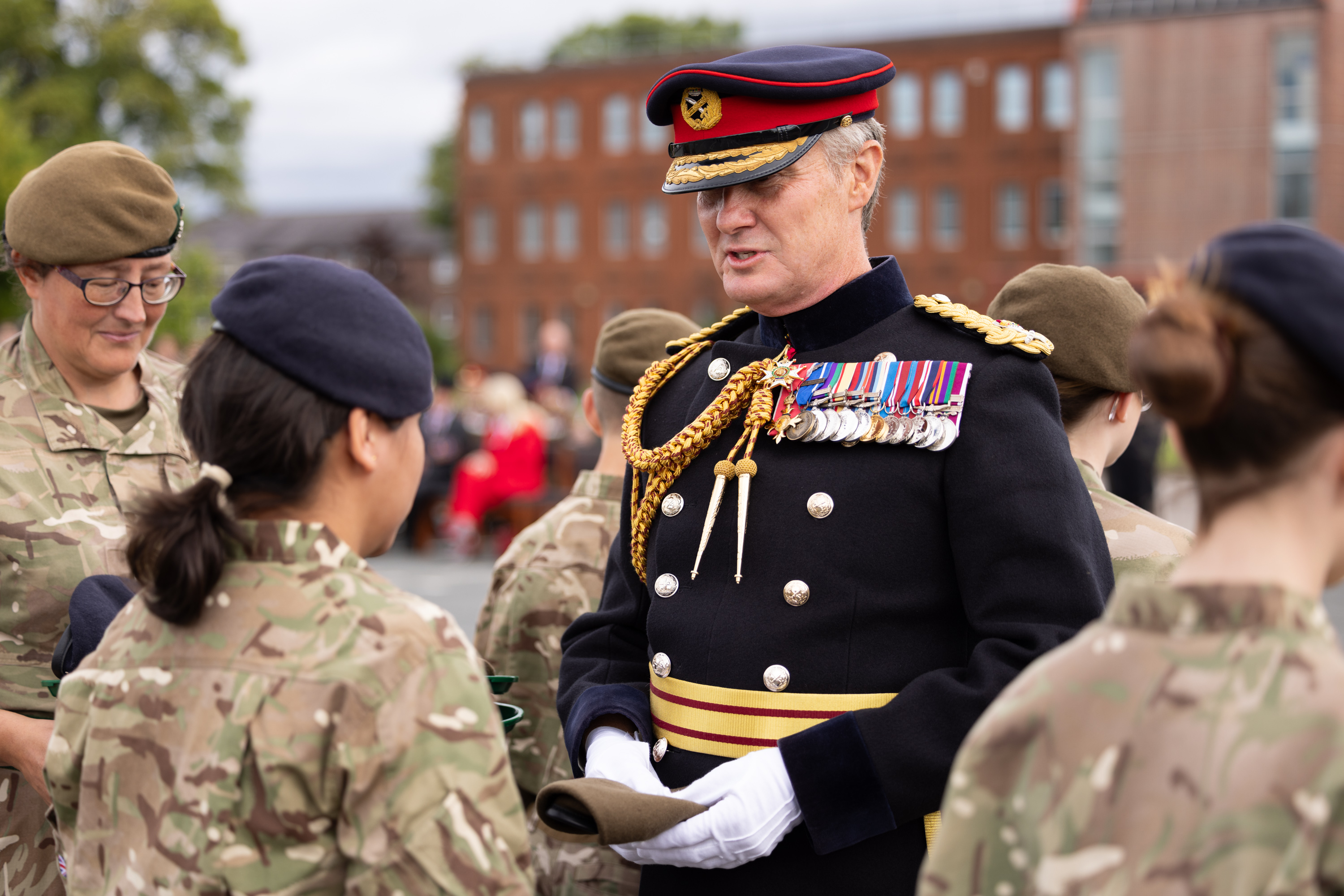 A person in a decorated military uniform, featuring numerous medals and a hat with a red and blue band, interacts with individuals in camouflage uniforms. They are outdoors with a building and trees in the background.