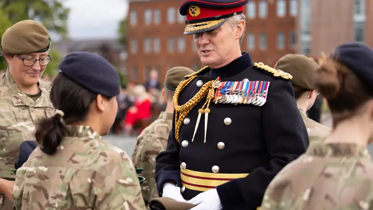 A person in a decorated military uniform, featuring numerous medals and a hat with a red and blue band, interacts with individuals in camouflage uniforms. They are outdoors with a building and trees in the background.