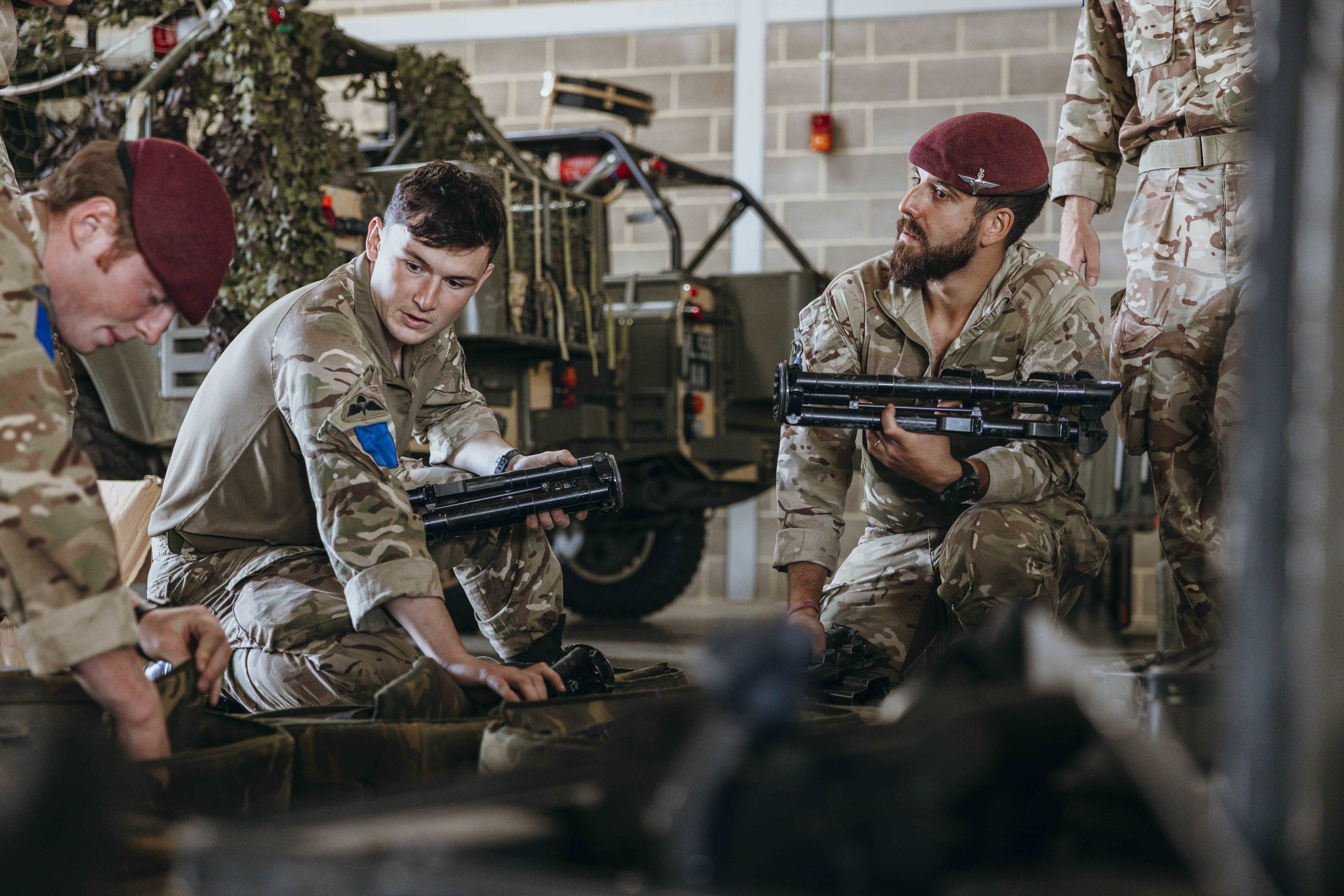Soldiers in camouflage uniforms and maroon berets sitting indoors, handling military equipment near a camouflaged vehicle.