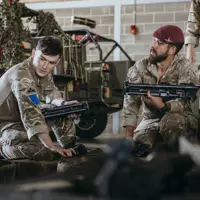 Soldiers in camouflage uniforms and maroon berets sitting indoors, handling military equipment near a camouflaged vehicle.