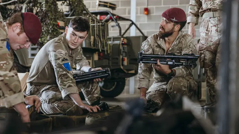 Soldiers in camouflage uniforms and maroon berets sitting indoors, handling military equipment near a camouflaged vehicle.