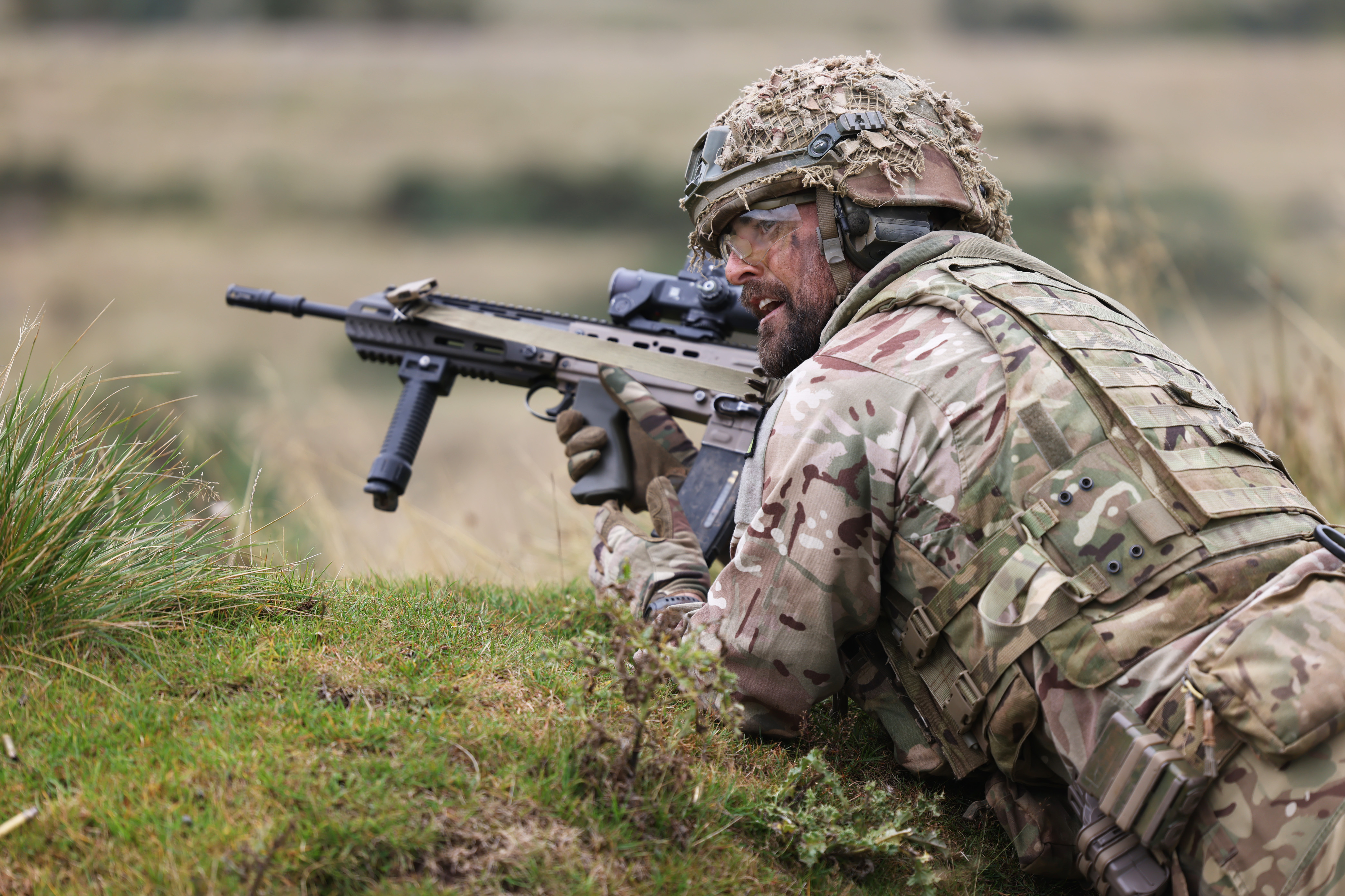 Soldier in camouflage gear aiming a rifle while lying prone in a grassy field during training or combat.