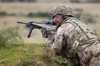 Soldier in camouflage gear aiming a rifle while lying prone in a grassy field during training or combat.