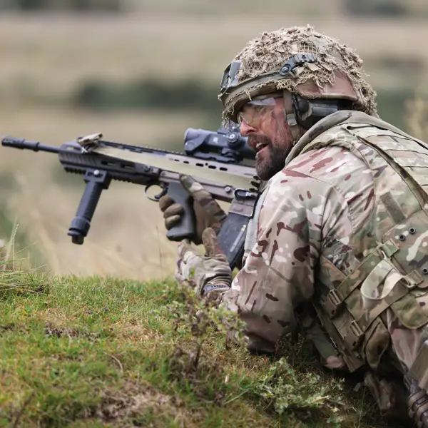 Soldier in camouflage gear aiming a rifle while lying prone in a grassy field during training or combat.