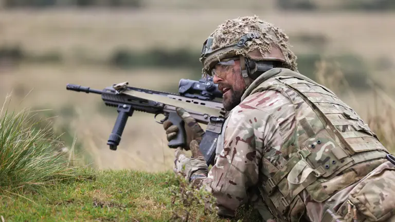 Soldier in camouflage gear aiming a rifle while lying prone in a grassy field during training or combat.