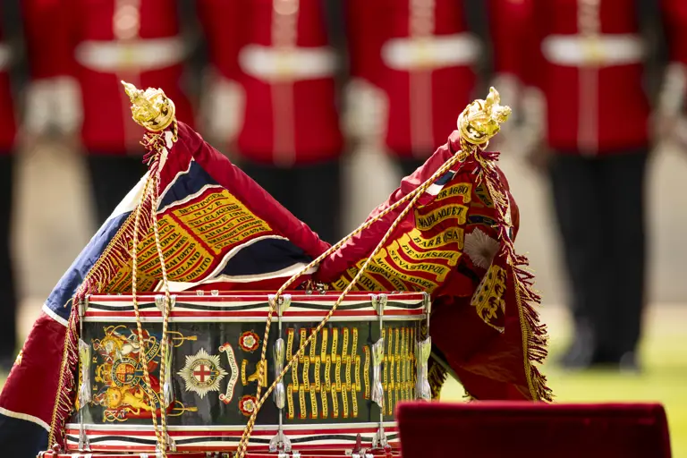 Close-up of a ceremonial military drum adorned with colorful battle honors and two ornate flags draped over it.