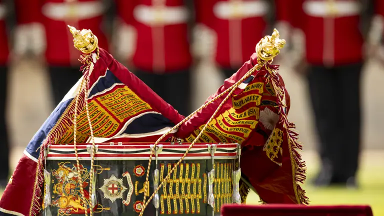 Close-up of a ceremonial military drum adorned with colorful battle honors and two ornate flags draped over it.