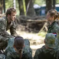A person in camouflage clothing crouches and speaks to another person dressed similarly, who is sitting opposite. They are surrounded by others in similar attire, listening attentively. The setting is outdoors, in a wooded area.