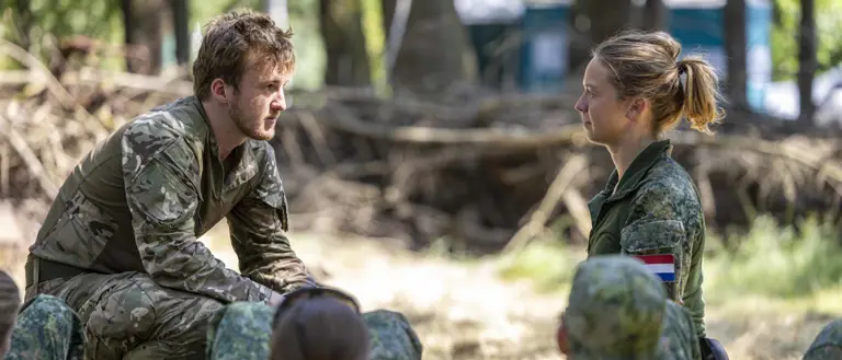 A person in camouflage clothing crouches and speaks to another person dressed similarly, who is sitting opposite. They are surrounded by others in similar attire, listening attentively. The setting is outdoors, in a wooded area.