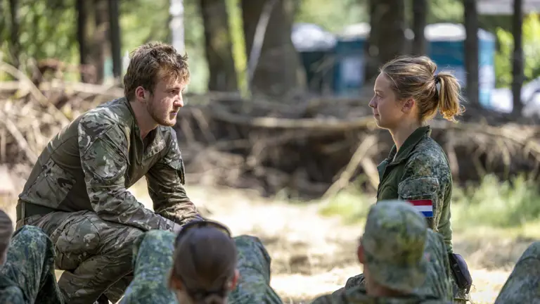 A person in camouflage clothing crouches and speaks to another person dressed similarly, who is sitting opposite. They are surrounded by others in similar attire, listening attentively. The setting is outdoors, in a wooded area.