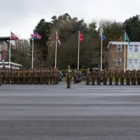 A uniformed officer with medals and a peaked cap converses with soldiers in brown uniforms and hats, set against a backdrop of trees.