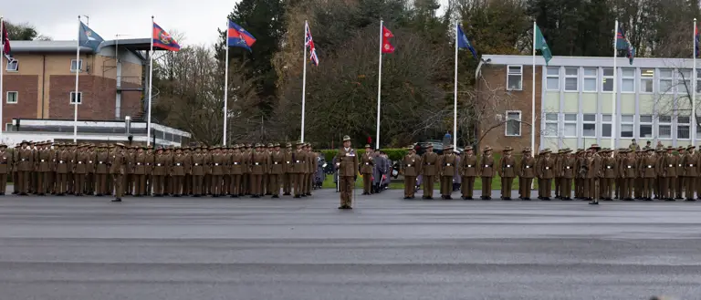 A uniformed officer with medals and a peaked cap converses with soldiers in brown uniforms and hats, set against a backdrop of trees.
