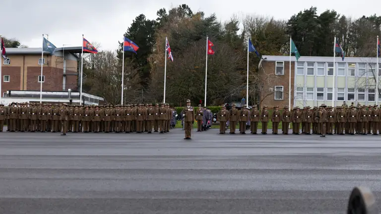 A uniformed officer with medals and a peaked cap converses with soldiers in brown uniforms and hats, set against a backdrop of trees.