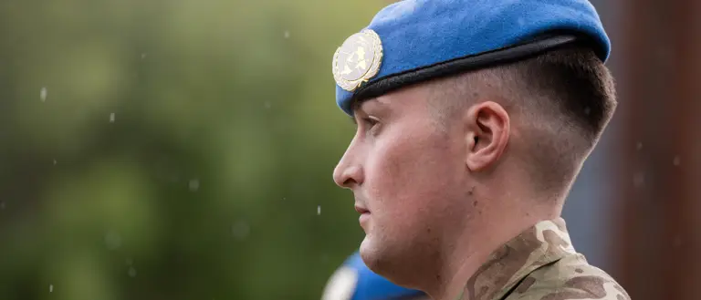 A soldier stands with a blue beret on his head.