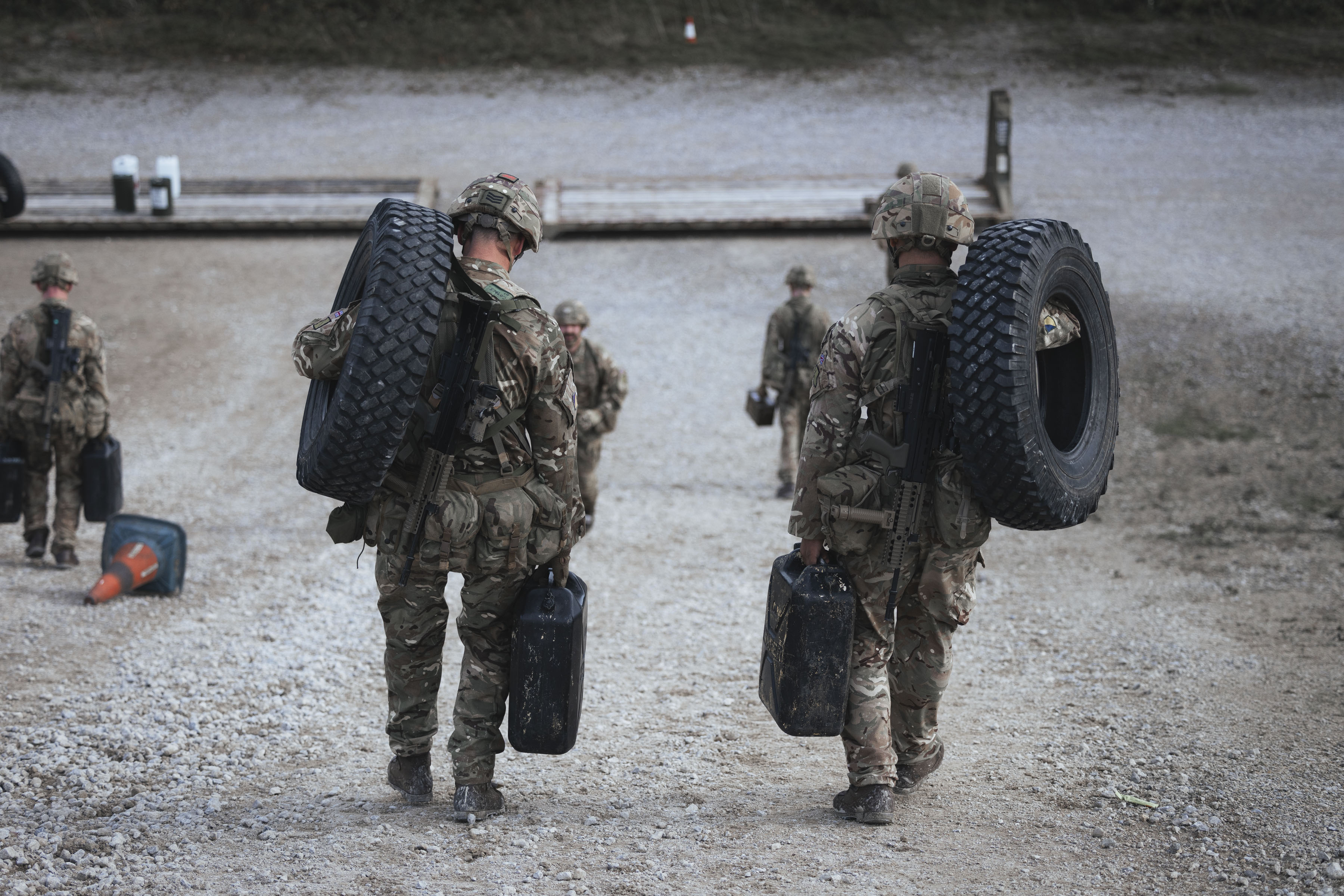 Two soldiers in full camouflage gear carry tyres and jerry cans during a training exercise.