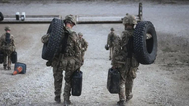 Two soldiers in full camouflage gear carry tyres and jerry cans during a training exercise.