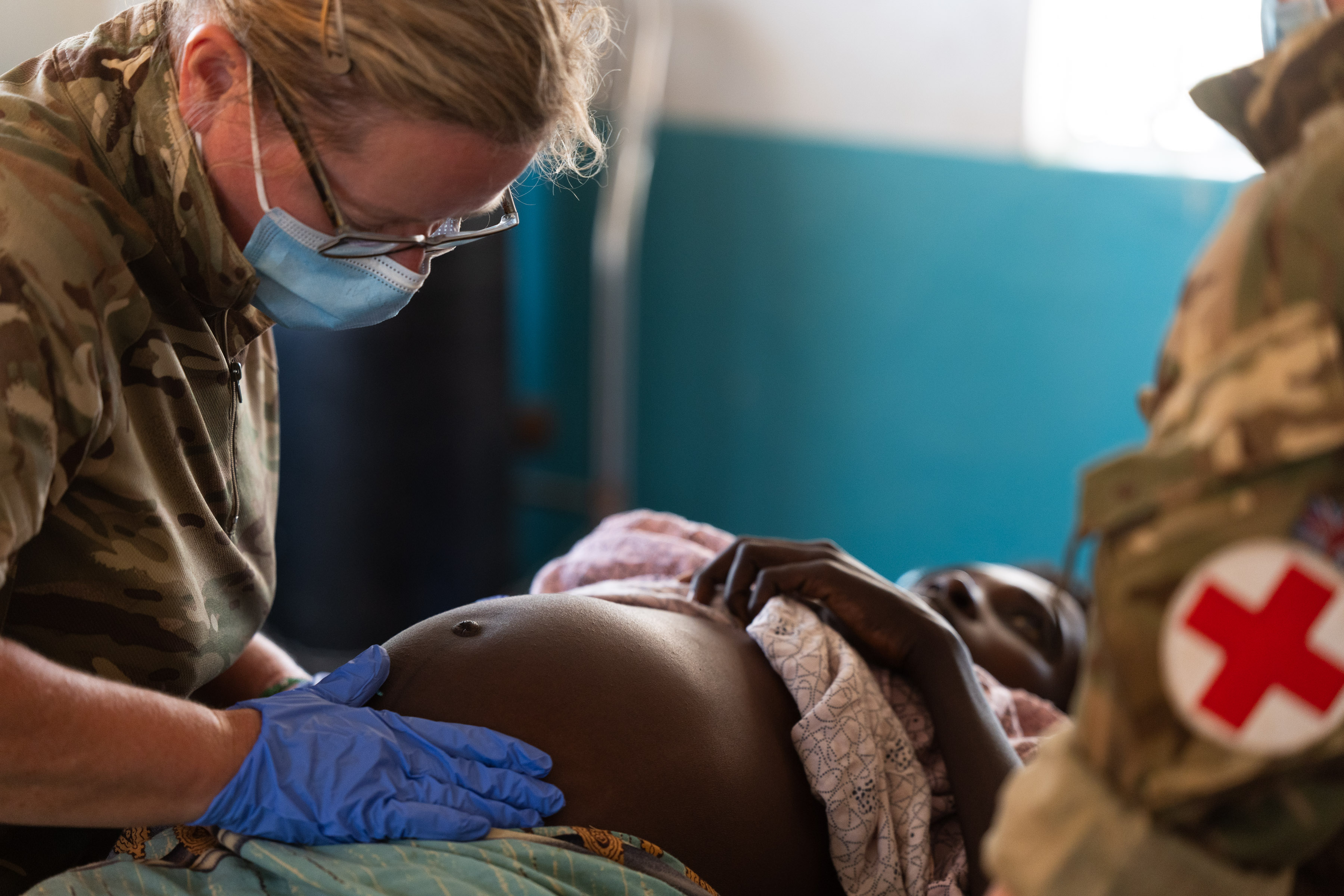 A healthcare worker in camouflage and a surgical mask examines a patient's abdomen in a medical setting.