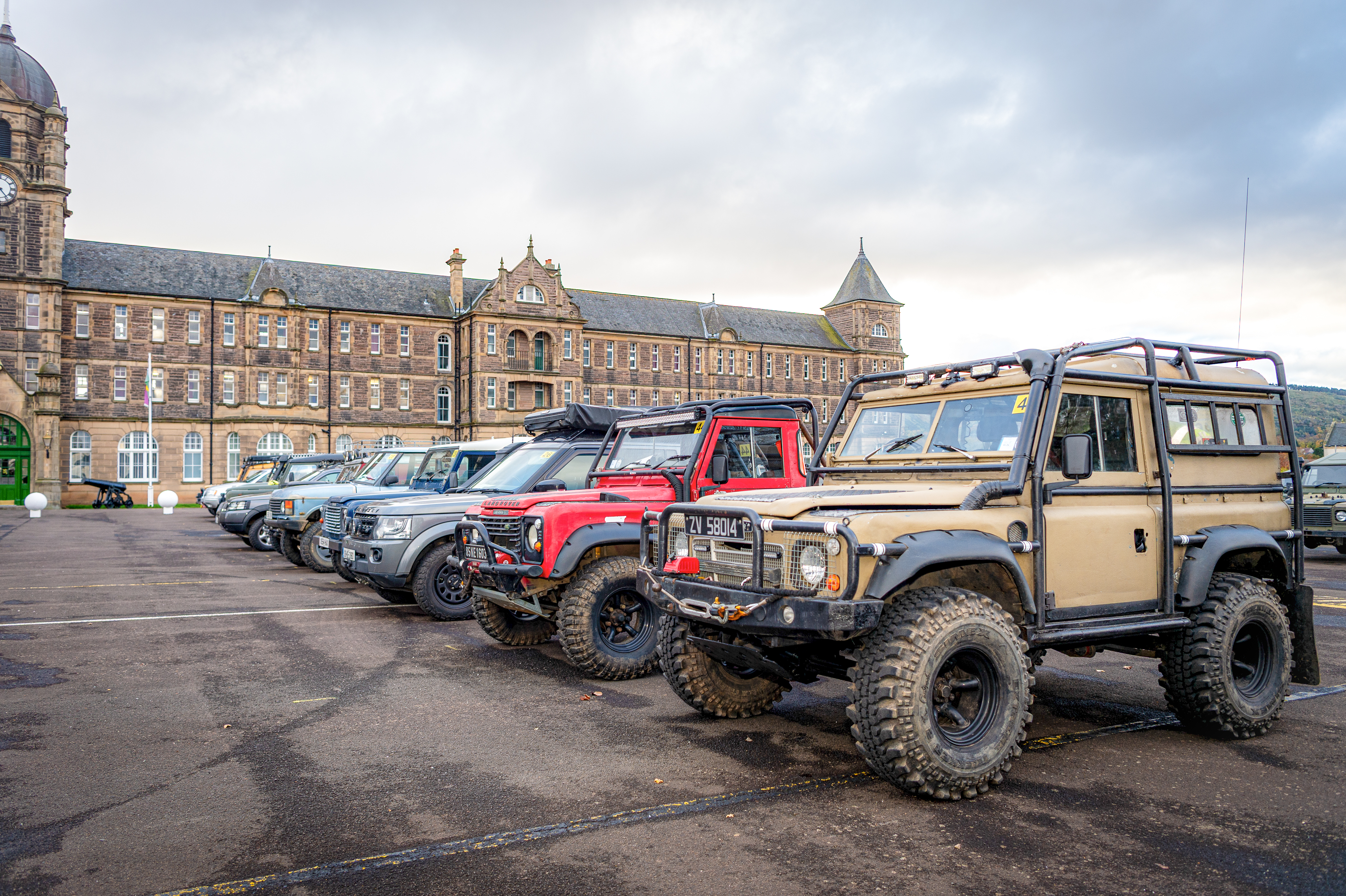 Line of rugged off-road vehicles parked on a wet street in front of a historic stone building with towers under an overcast sky.