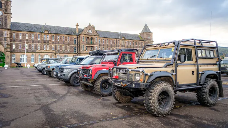 Line of rugged off-road vehicles parked on a wet street in front of a historic stone building with towers under an overcast sky.