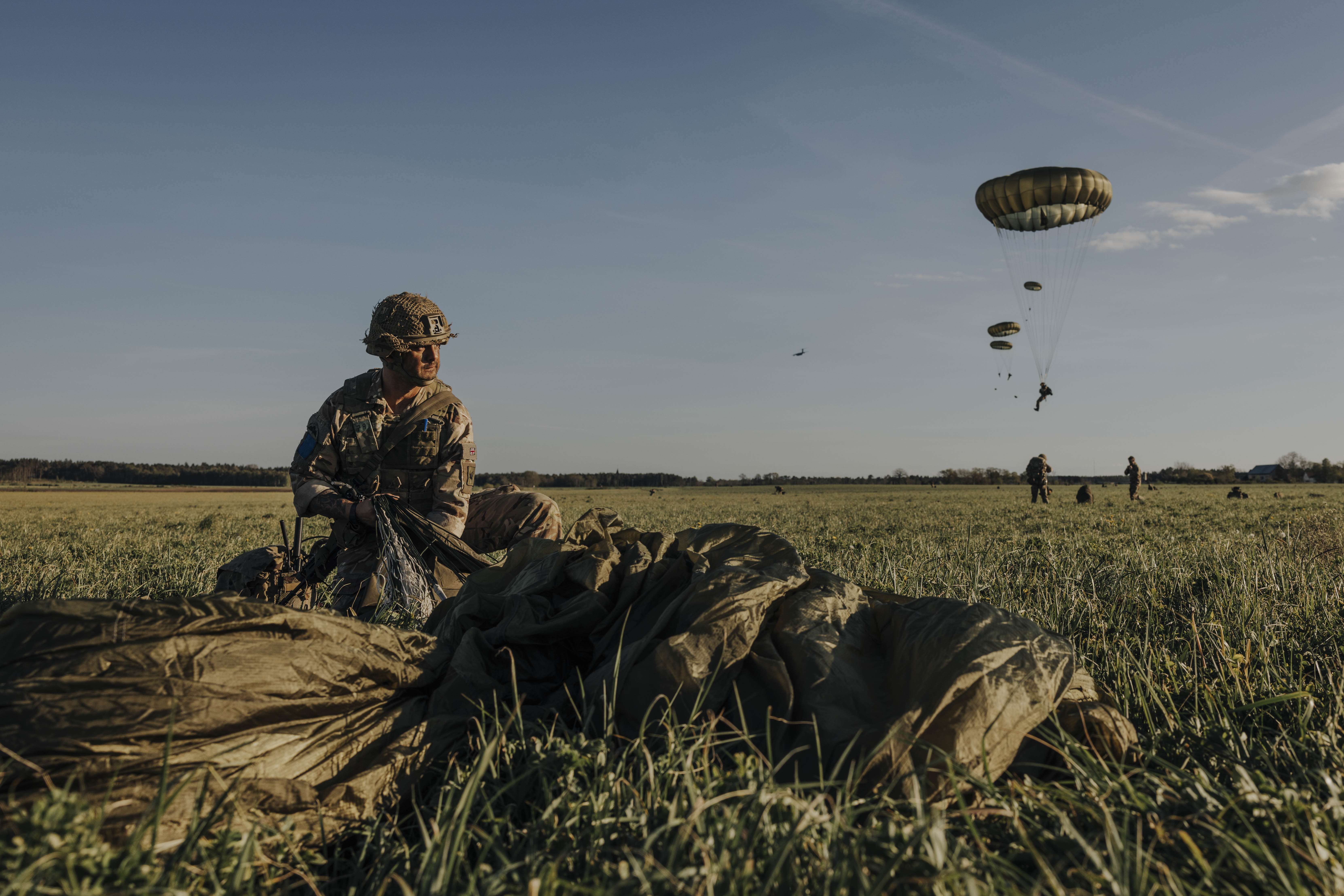 Paratrooper in combat camouflage uniform and helmet packs away his parachute after jumping from a United States A400 aircraft. The air craft is seen in the sky in the distance with other paratroopers in mid jump in the distance. 