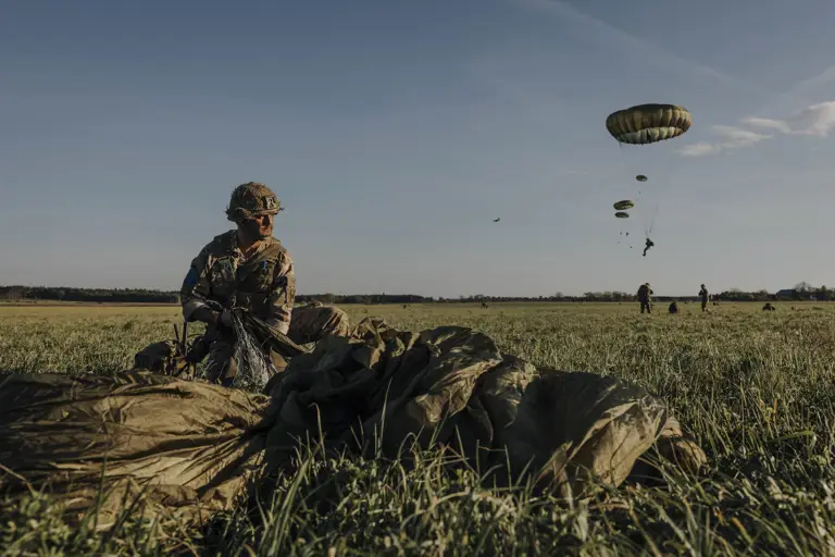 Paratrooper in combat camouflage uniform and helmet packs away his parachute after jumping from a United States A400 aircraft. The air craft is seen in the sky in the distance with other paratroopers in mid jump in the distance. 