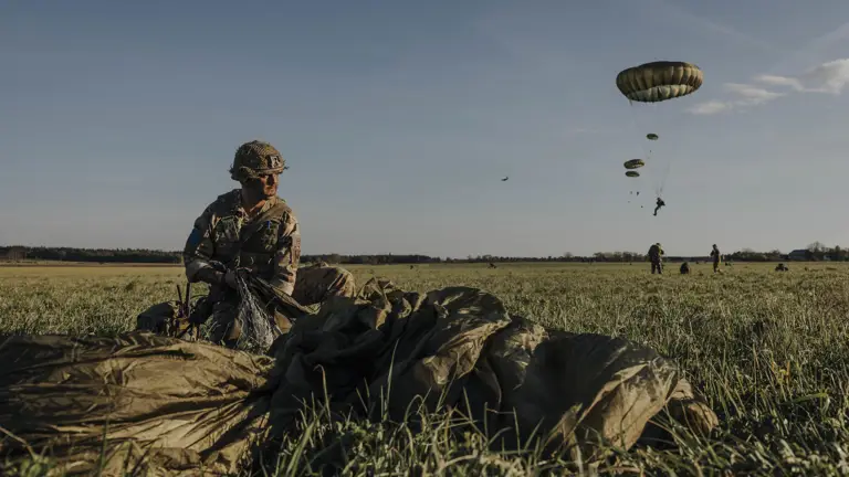 Paratrooper in combat camouflage uniform and helmet packs away his parachute after jumping from a United States A400 aircraft. The air craft is seen in the sky in the distance with other paratroopers in mid jump in the distance.