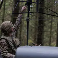 Soldier in camouflage uniform adjusting a large antenna mounted on a vehicle in a dense forest setting.