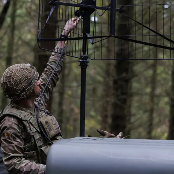 Soldier in camouflage uniform adjusting a large antenna mounted on a vehicle in a dense forest setting.