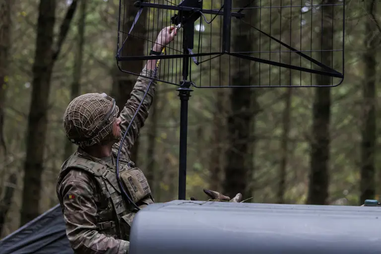 Soldier in camouflage uniform adjusting a large antenna mounted on a vehicle in a dense forest setting.