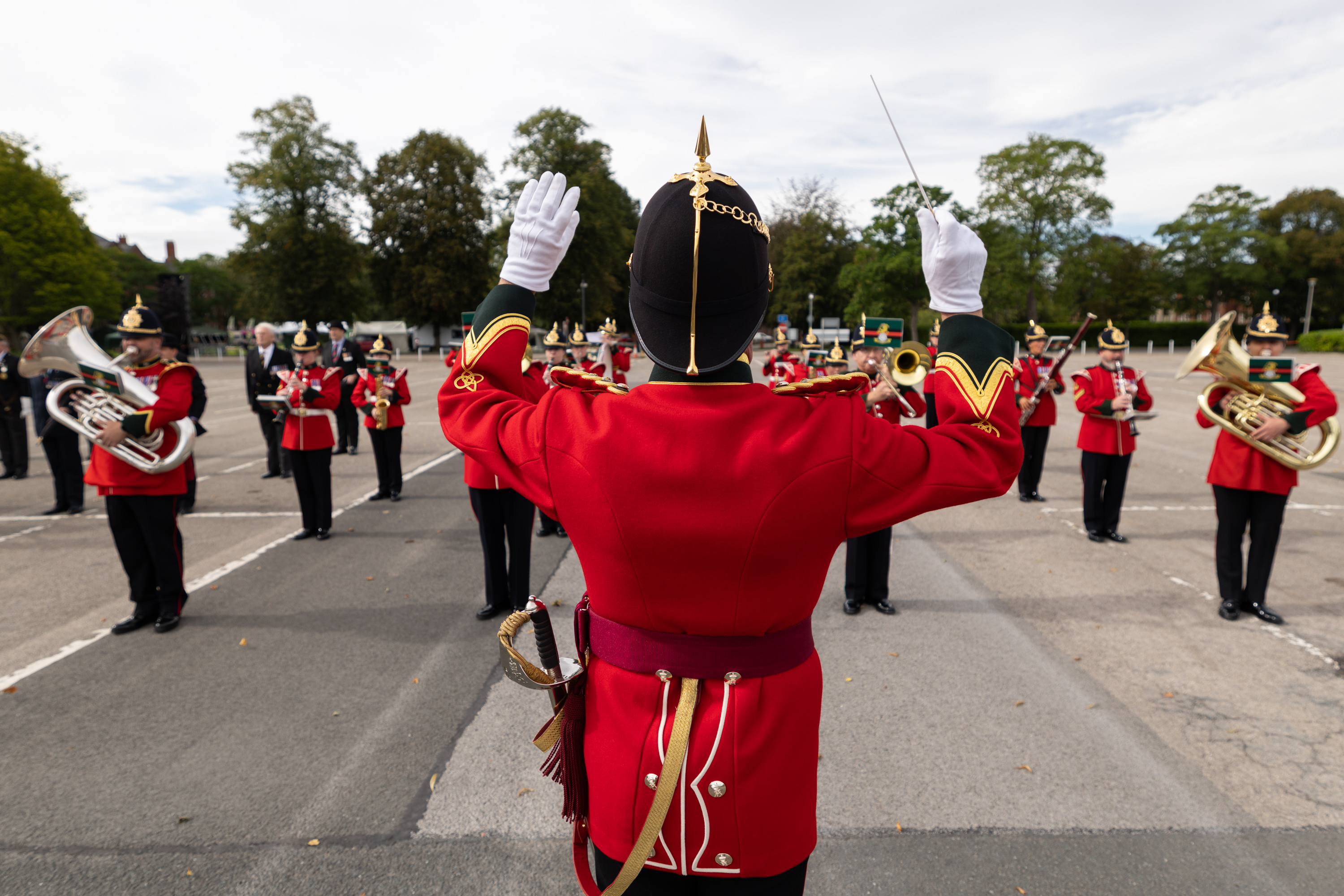 A marching band performs outdoors, directed by a person in a red uniform with black and gold details. The musicians, also in red, play brass instruments on a paved area surrounded by trees.