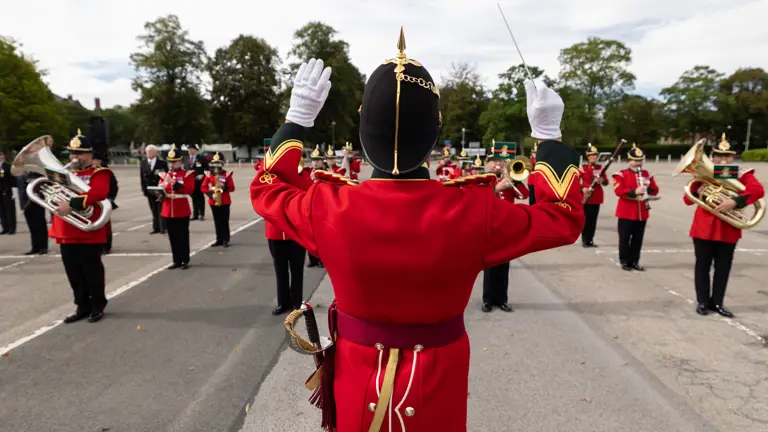 A marching band performs outdoors, directed by a person in a red uniform with black and gold details. The musicians, also in red, play brass instruments on a paved area surrounded by trees.