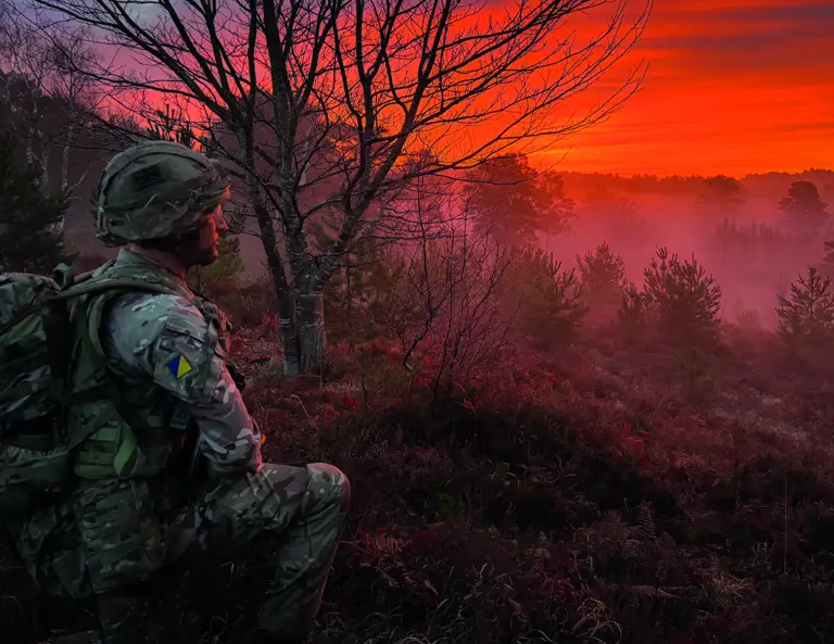 Royal Logistic Corps soldier knelt down wearing camouflage uniform, a helmet and a large green bag on his back. He's looking into the bright orange and pink sunset
