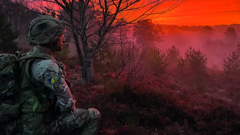 Royal Logistic Corps soldier knelt down wearing camouflage uniform, a helmet and a large green bag on his back. He's looking into the bright orange and pink sunset