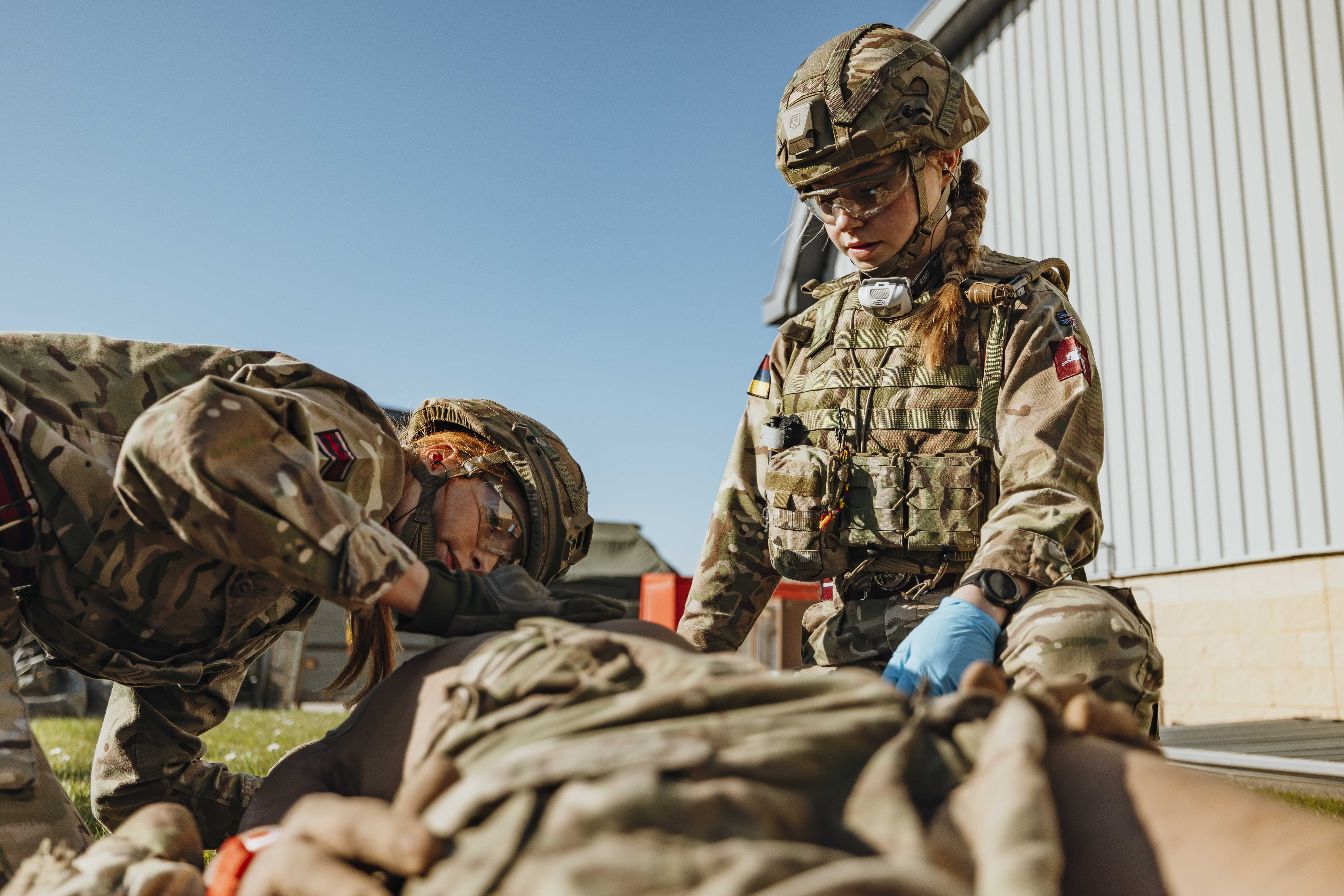 Two soldiers in camouflage uniforms provide medical aid to a comrade lying on the ground outdoors near a building.