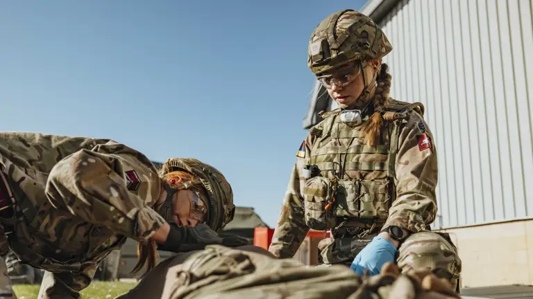 Two soldiers in camouflage uniforms provide medical aid to a comrade lying on the ground outdoors near a building.