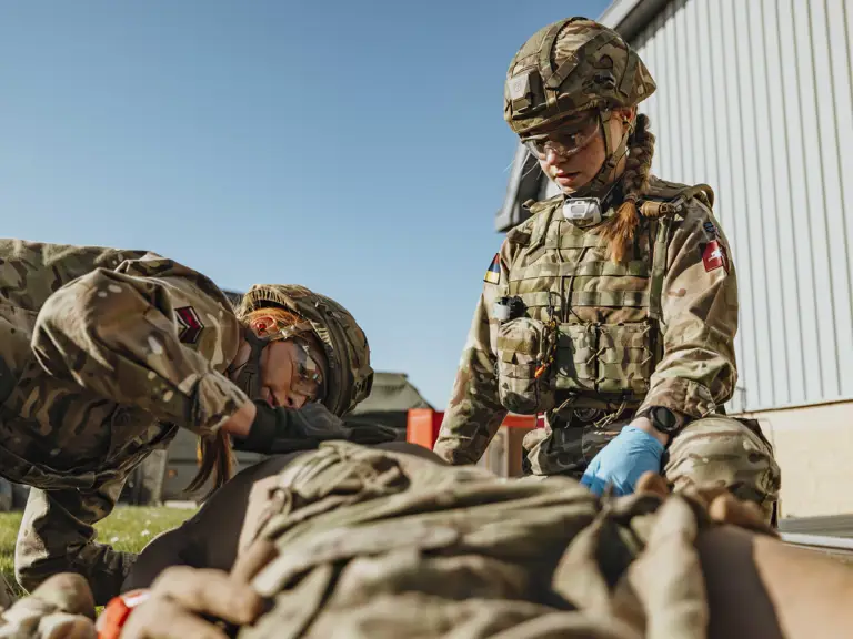 Two soldiers in camouflage uniforms provide medical aid to a comrade lying on the ground outdoors near a building.