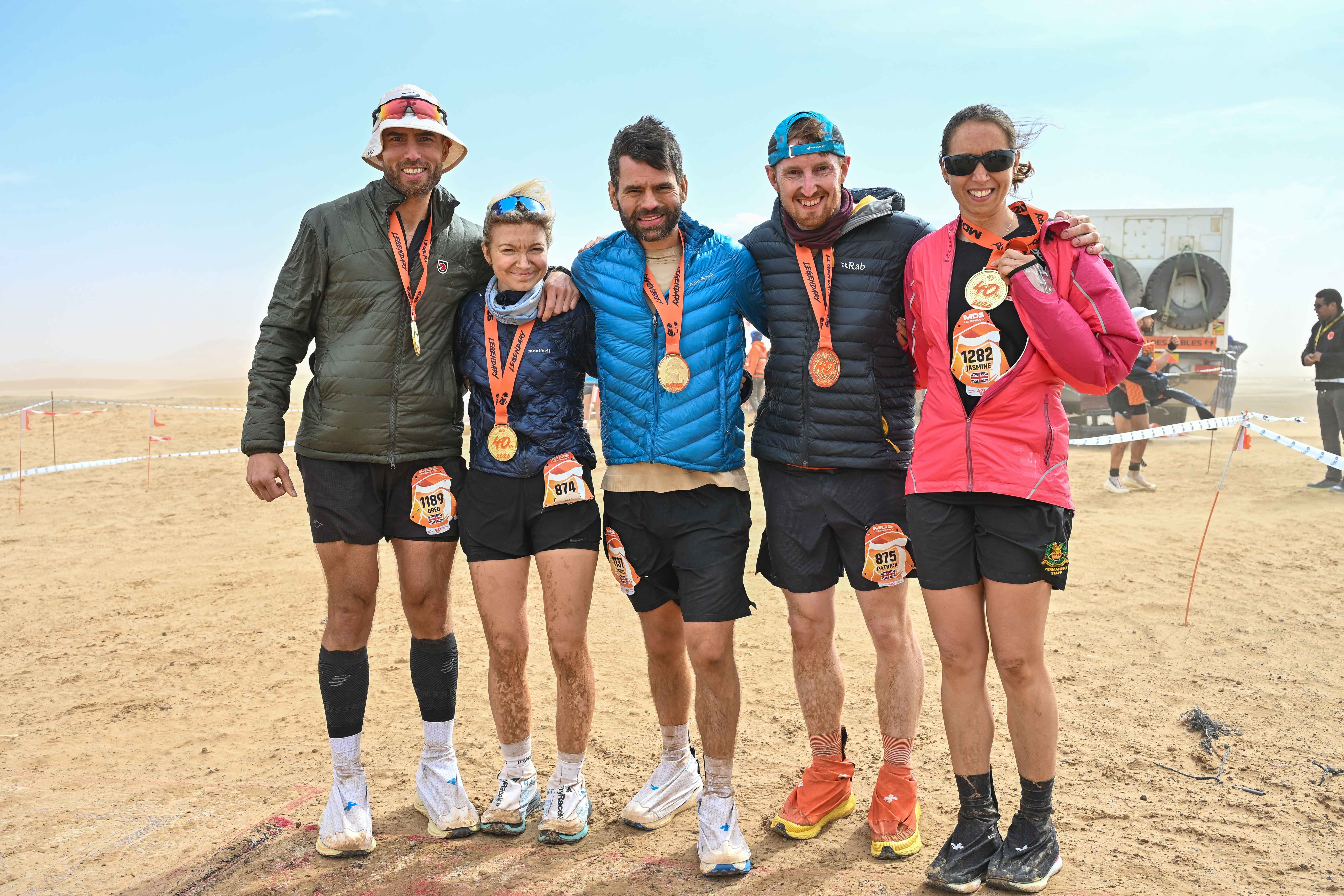 Five people stood in running gear and coats with medals hanging from their necks in a desert background.