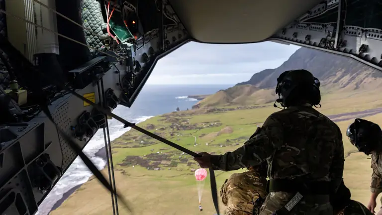 Inside a helicopter, soldiers in camouflage gear prepare for a drop over a coastal landscape with cliffs and fields. The scene is dynamic and intense.