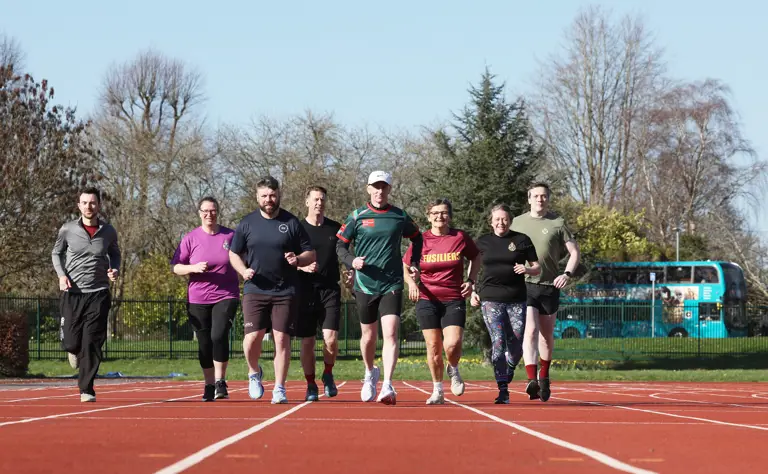 Seven runners on a track, smiling as they jog side-by-side. They display a sense of camaraderie under a clear blue sky, with trees in the background.