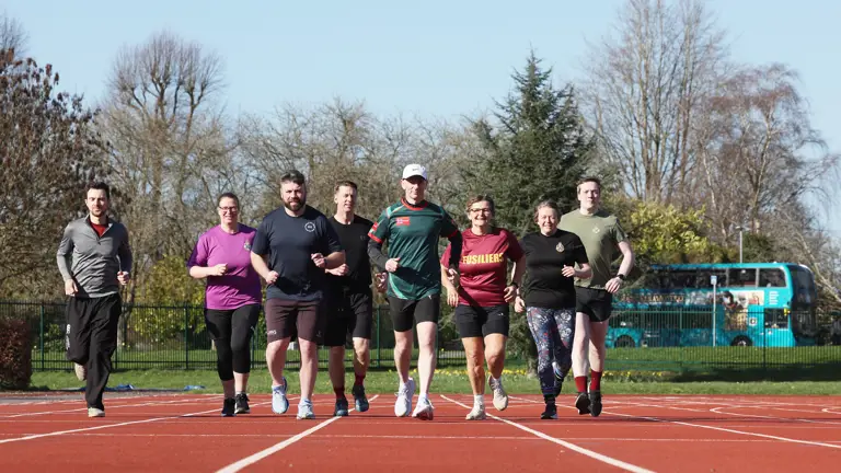 Seven runners on a track, smiling as they jog side-by-side. They display a sense of camaraderie under a clear blue sky, with trees in the background.
