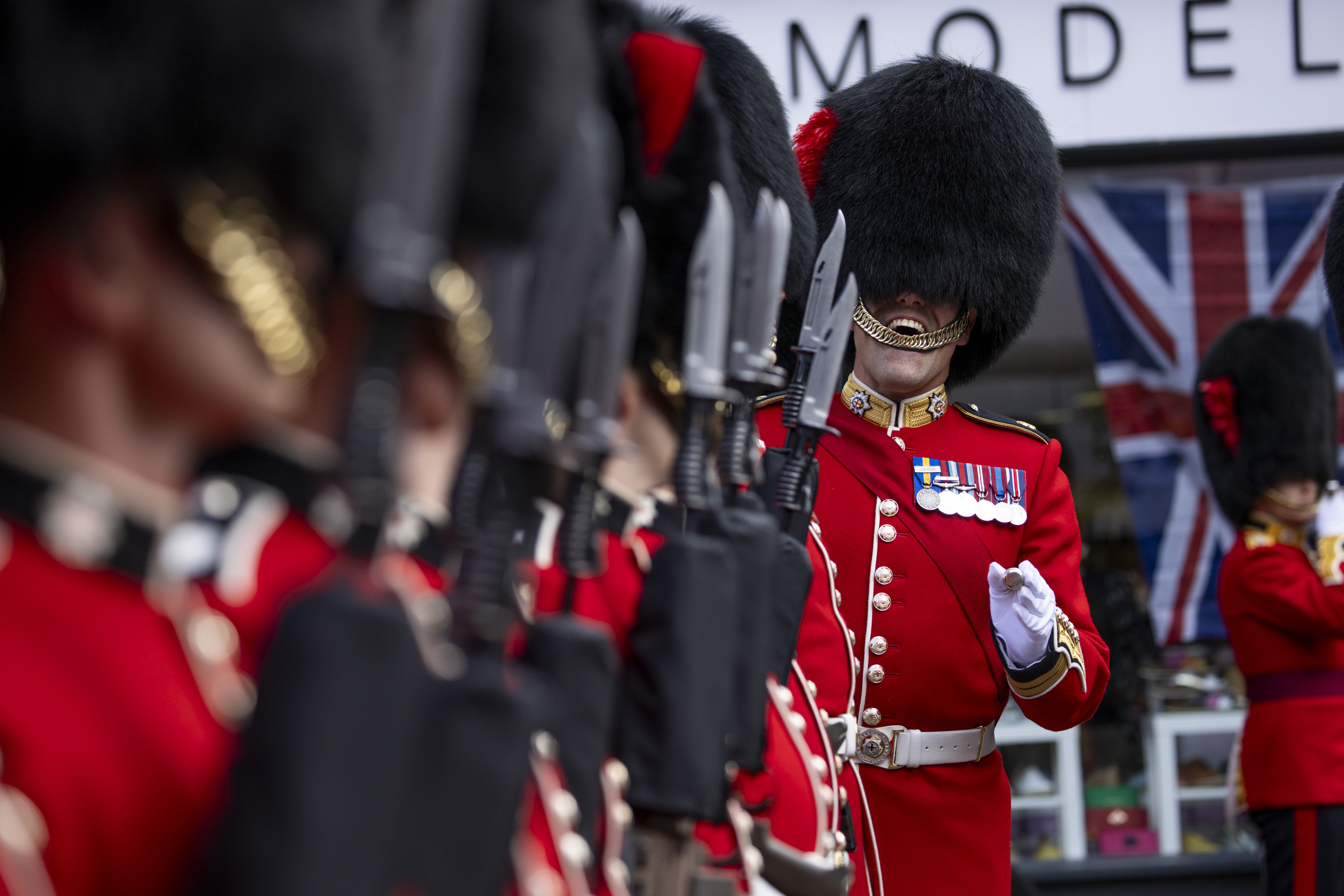 A soldier ensures soldiers wearing red tunics and black bearskin hats are lined up straight.