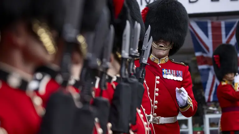 A soldier ensures soldiers wearing red tunics and black bearskin hats are lined up straight.